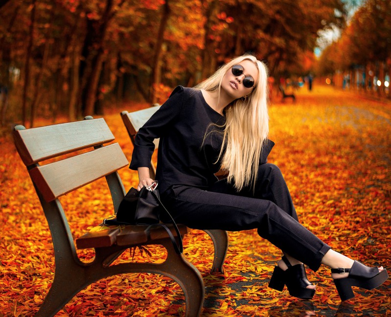 A blonde girl in all-black assemble sitting on the bench in the park in autumn